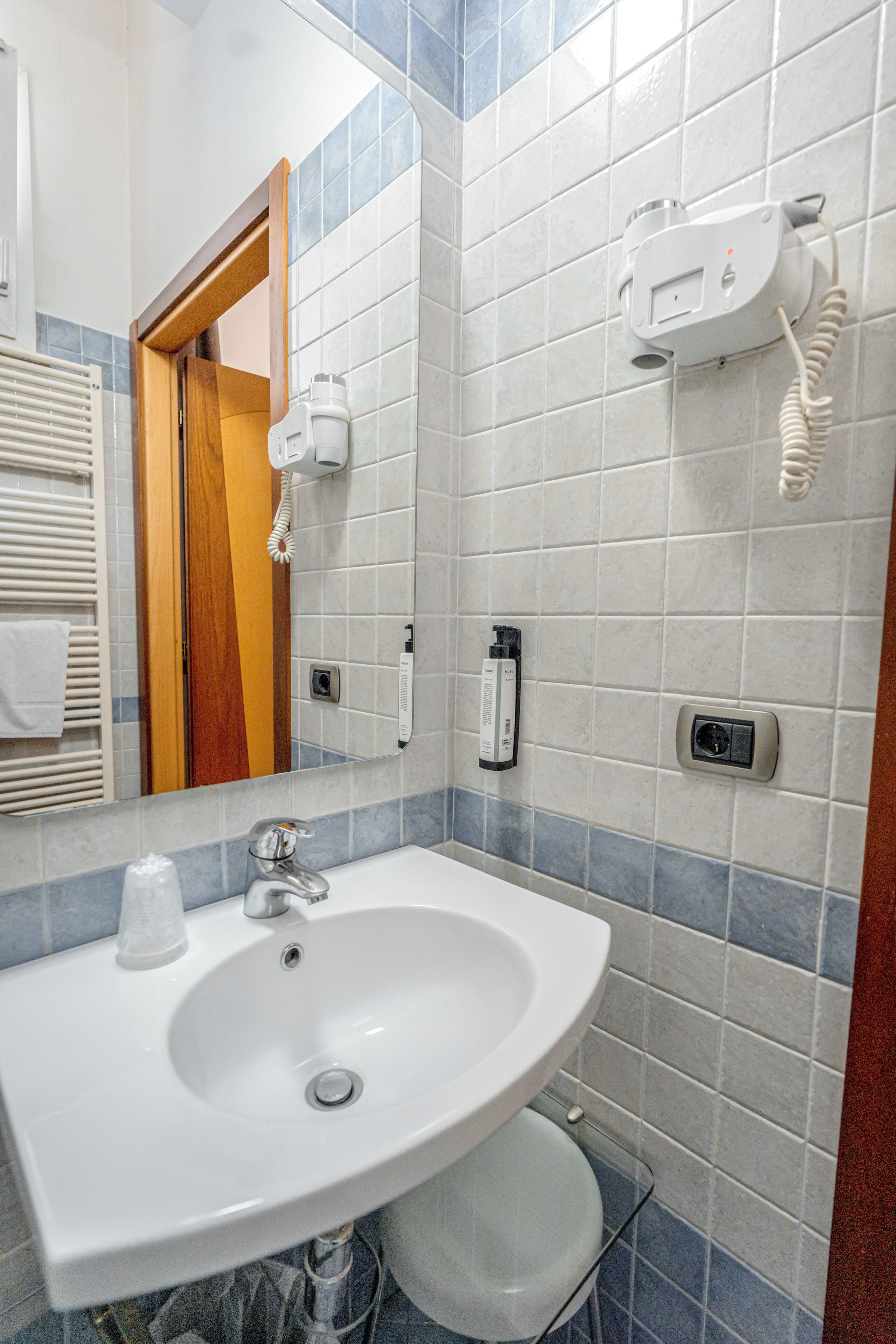 Un baño con lavamanos blanco, espejo, toallero y secador de pelo montado en la pared.