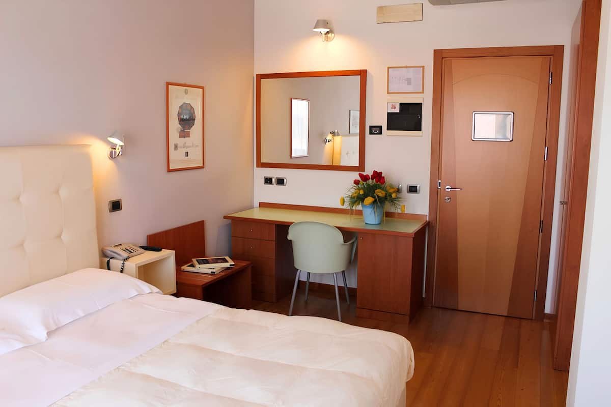 Une chambre d’hôtel avec un lit, un bureau, une chaise, une porte et un miroir fixé au mur.