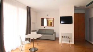 A modern living room with a sofa, a dining table, a TV mounted on the wall, and a small table. - Rubicone Hotel (Savignano sul Rubicone)