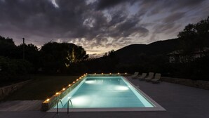 Piscina rectangular con borde de piedra y luces a lo largo del borde, rodeada por una solana empedrada y sillones de descanso.