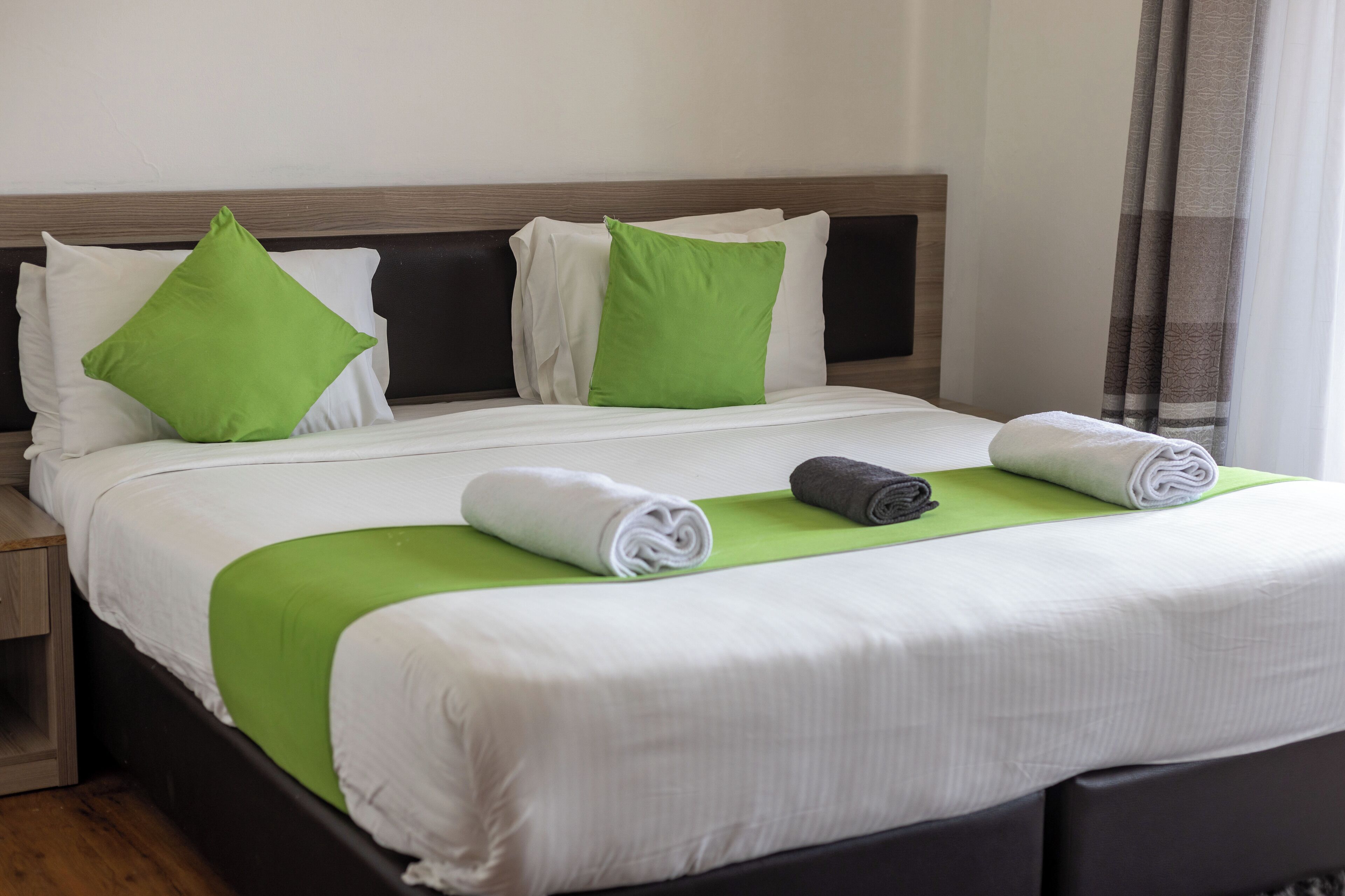 Una cama bien tendida con ropa de cama blanca y verde, un cabecero de madera y dos toallas enrolladas.