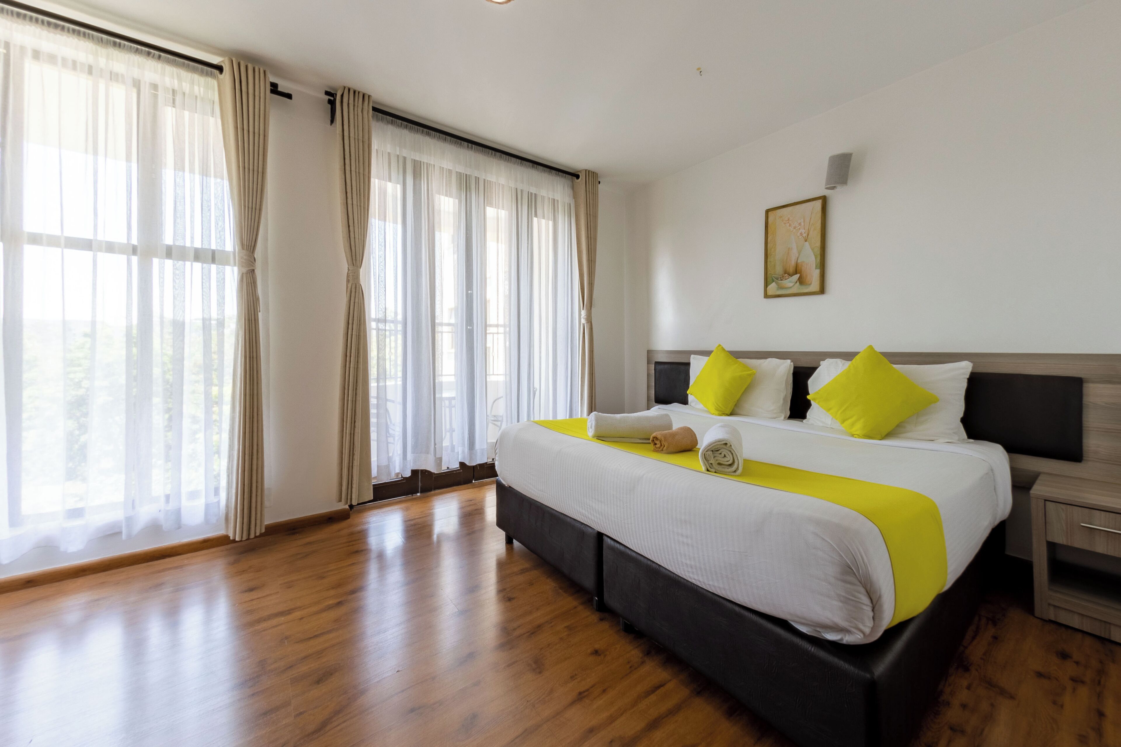 A hotel room with a large bed, wooden flooring, and a view of the outdoors through sheer curtains.