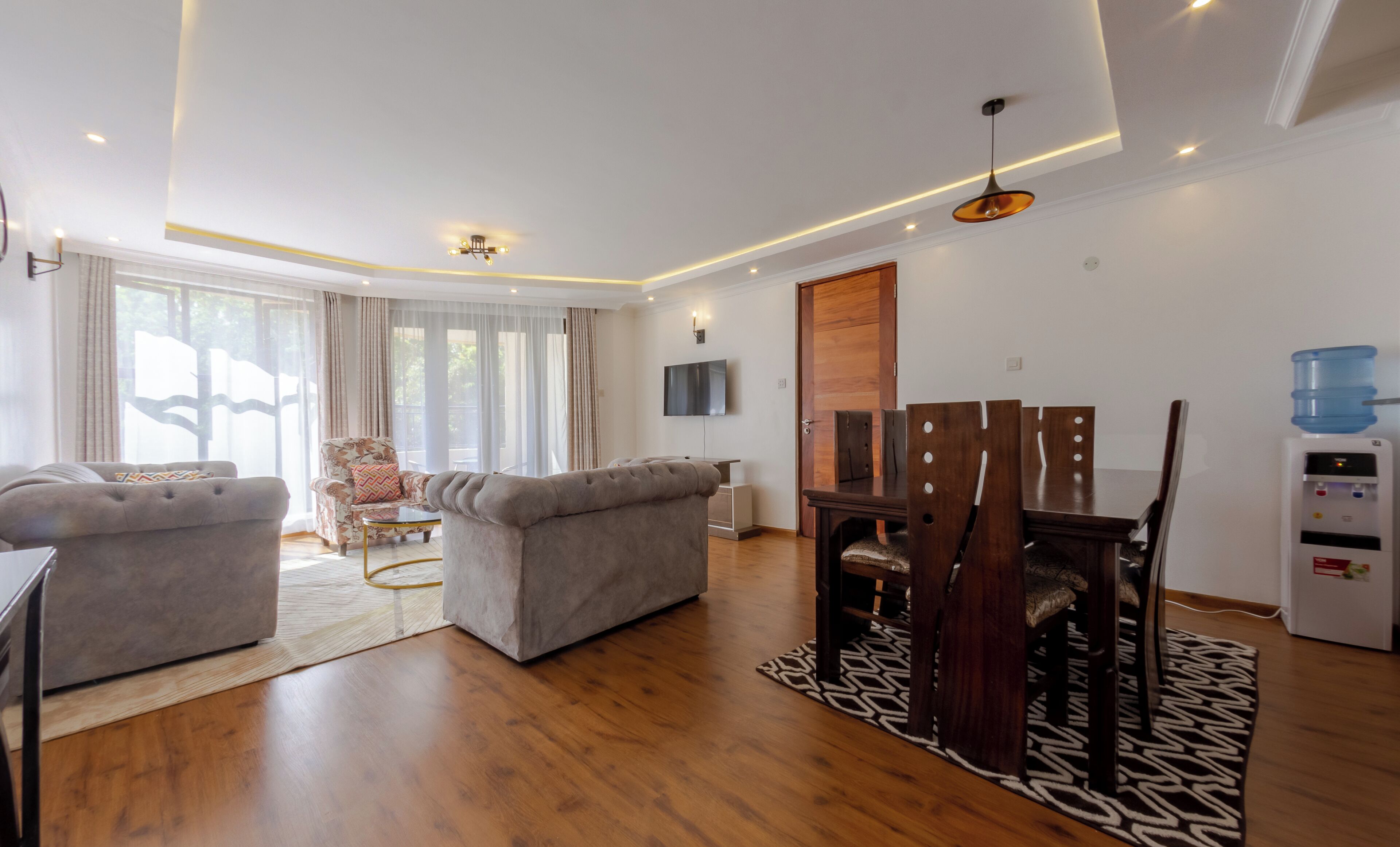 Amplia sala de estar con sofá, sillón y una mesa de comedor con sillas. Un televisor de pantalla plana está montado en la pared, y hay un dispensador de agua a la derecha.