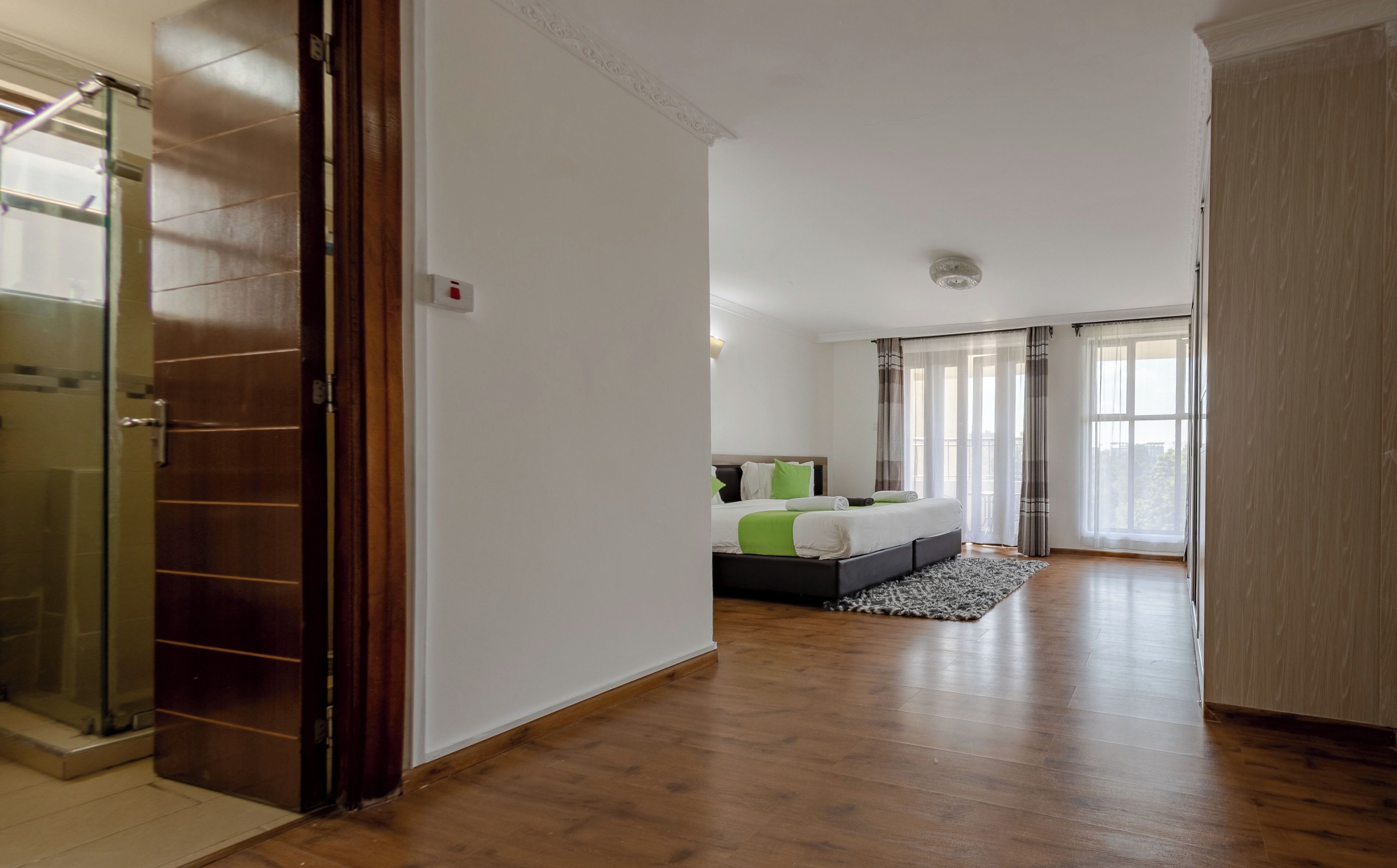 A hotel room with a wooden floor, a bed with green and white bedding, a small rug, and a view of the city through large windows.