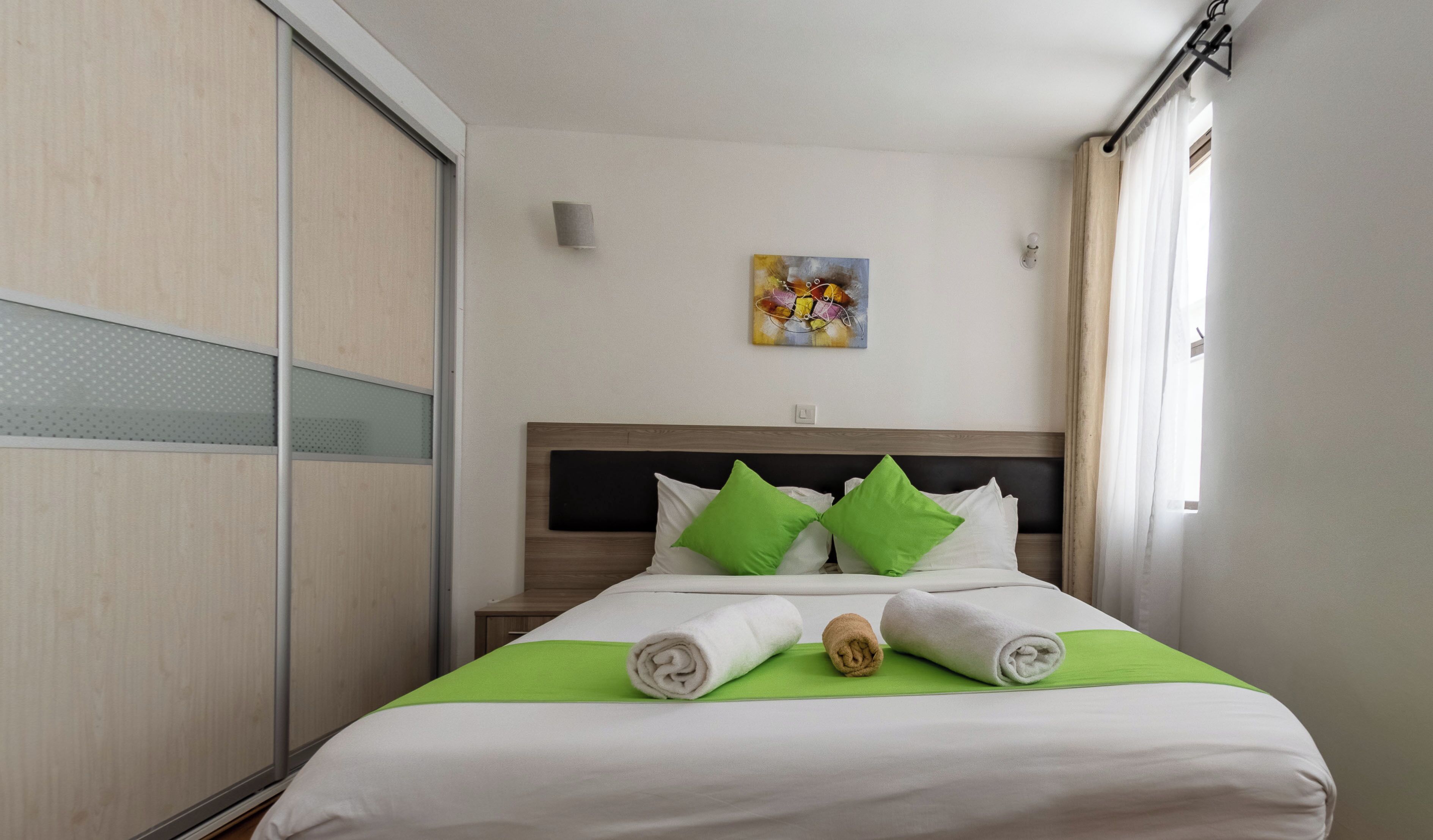 Hotelkamer met een bed, groene kussens, een schilderij aan de muur en een raam met gordijnen.