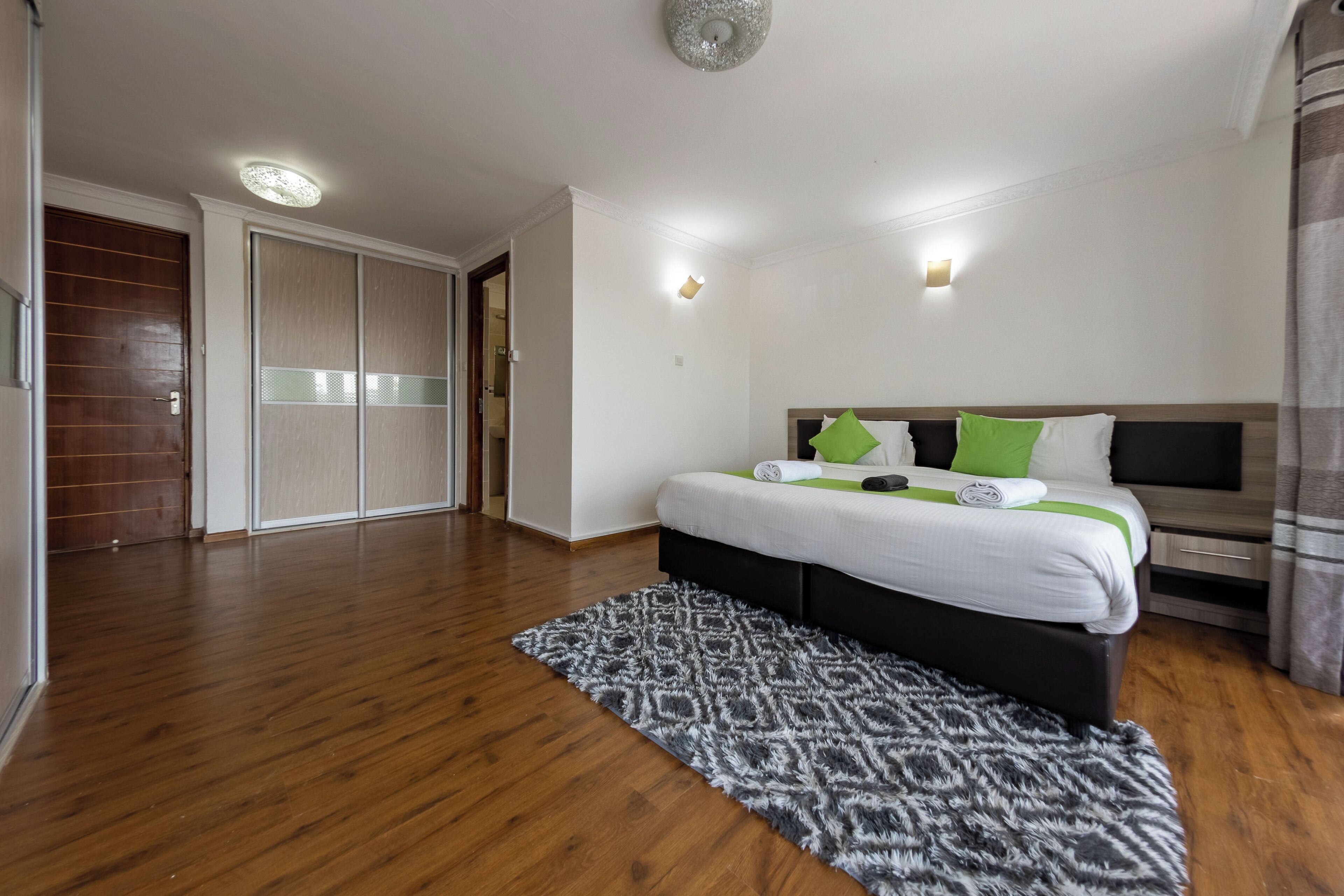 A bedroom with a wooden floor, a bed with white bedding and green pillows, a black headboard, and a grey patterned rug.