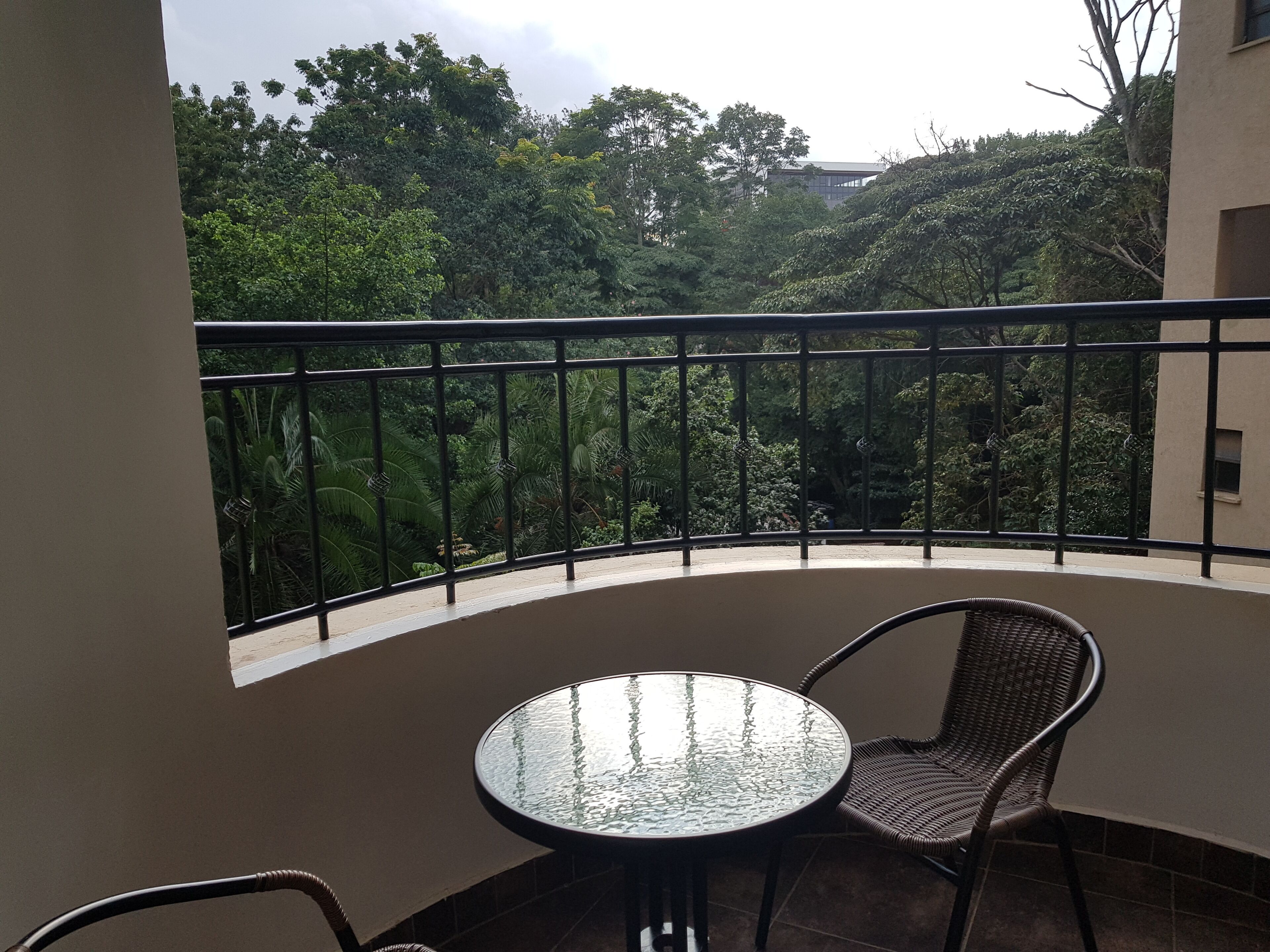 A balcony with a round table and two wicker chairs, overlooking a lush garden and a building in the background.