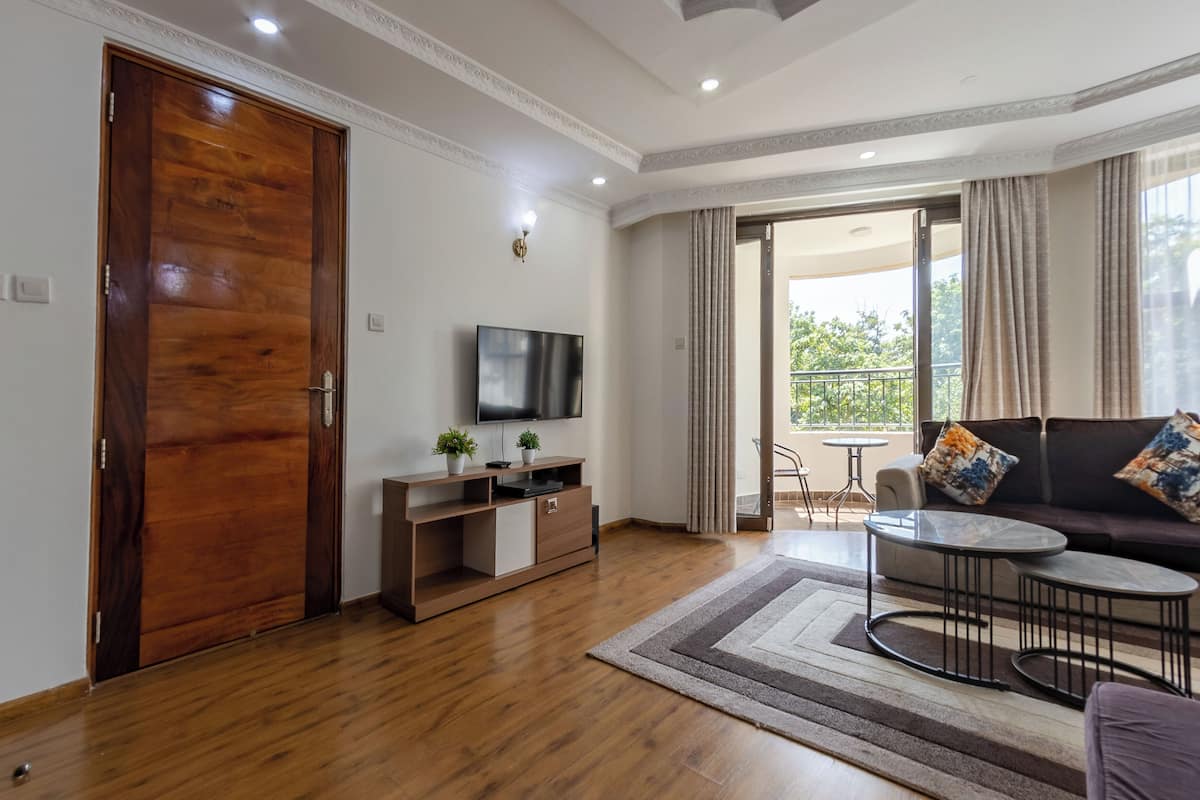 A modern living room with a wooden door, a flat-screen TV on a wooden console, a sofa with patterned cushions, a small round table, and a view of the outdoors through a balcony.