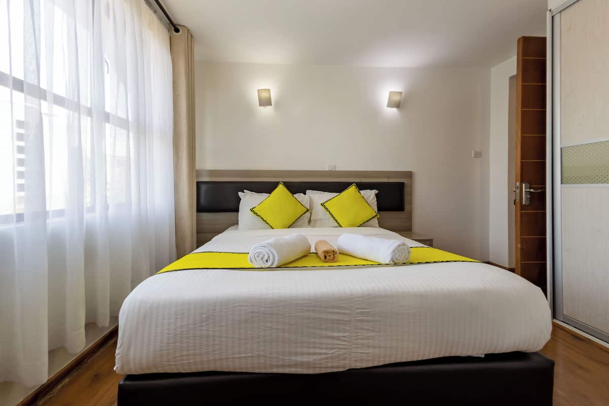 A neatly made bed with white bedding and yellow accents, a wooden headboard, and a bedside table with a lamp.