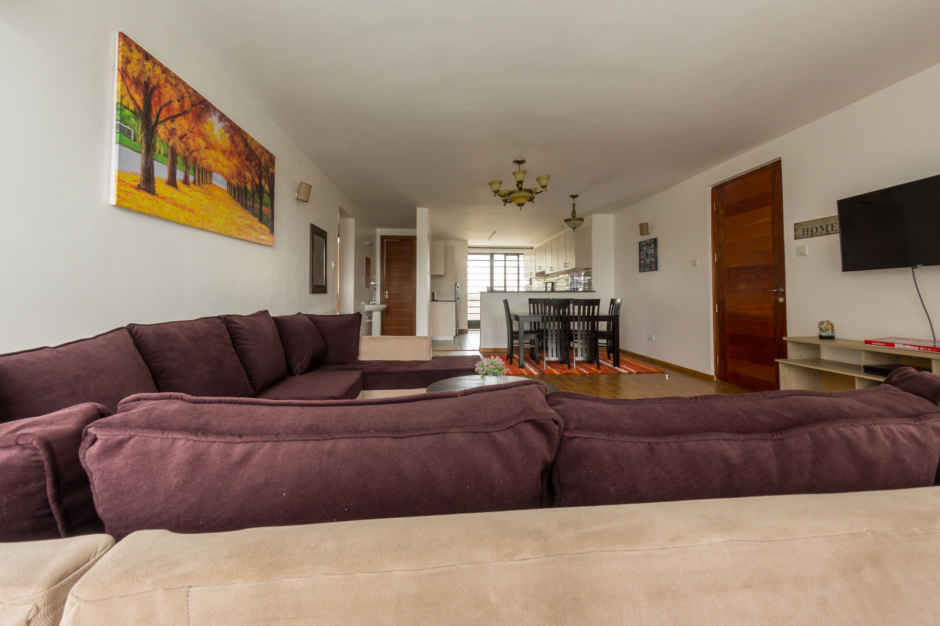 Uma sala de estar com um amplo sofá modular, uma área de jantar com mesa e cadeiras, uma cozinha ao fundo e uma TV fixada na parede.