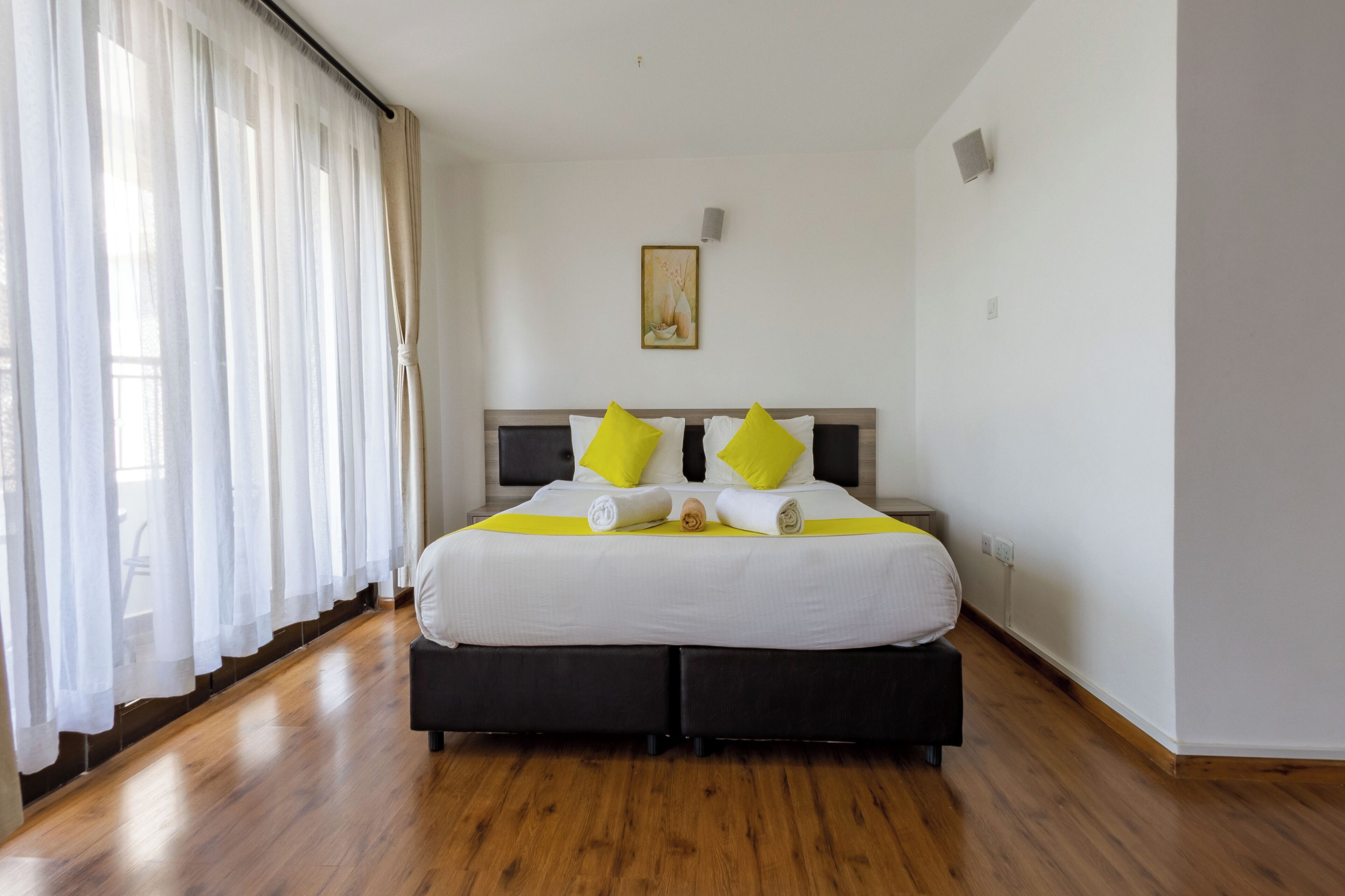 Una cama bien tendida con ropa de cama blanca y dos almohadas amarillo brillante, un piso de madera y una ventana con cortinas translúcidas.