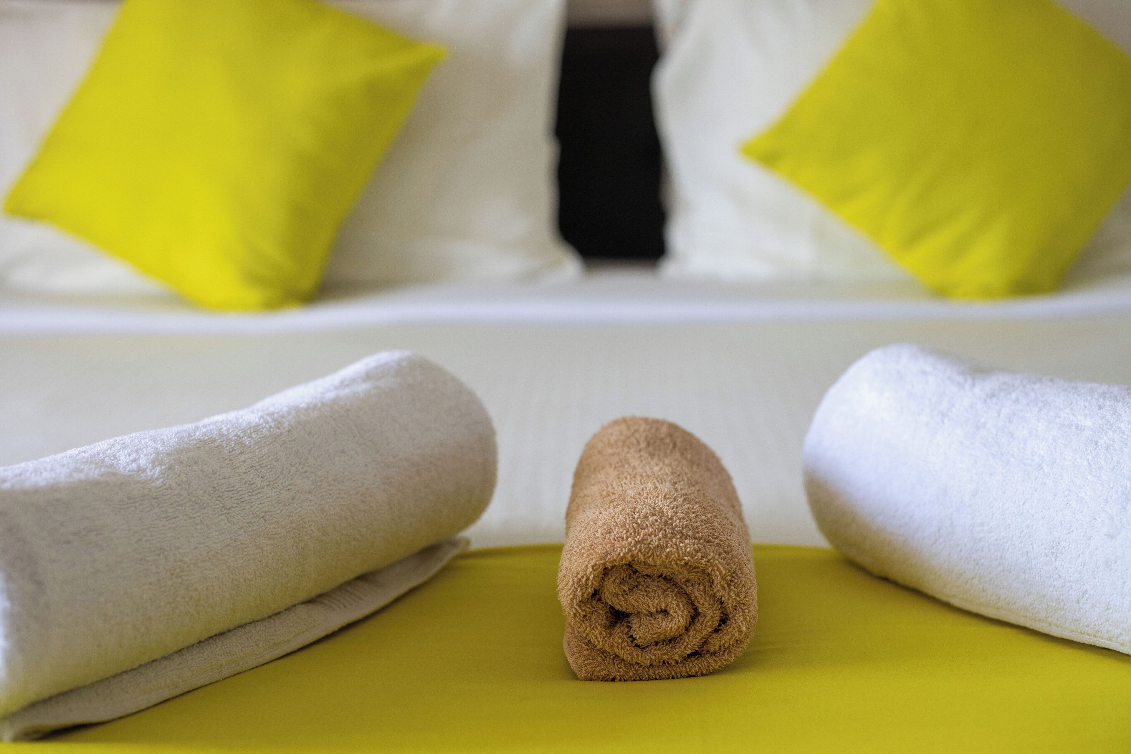Un lit bien fait, avec une literie blanche et deux oreillers, un jaune et un noir, ainsi que deux serviettes enroulées, une beige et une blanche, posées sur une surface jaune.