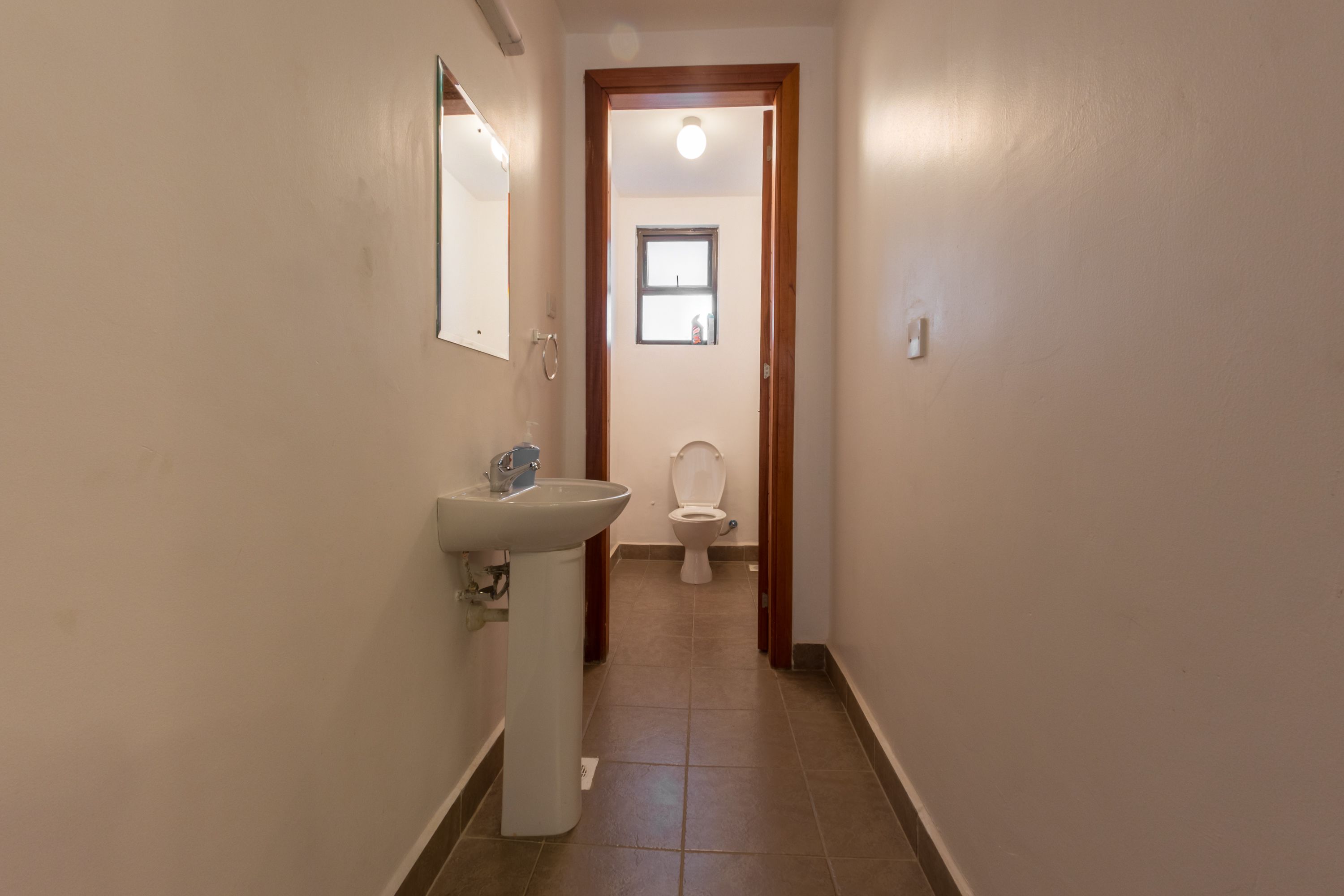 Een smalle gang met een wit wastafel en toilet, houten deurkozijn en een klein raam aan het einde.