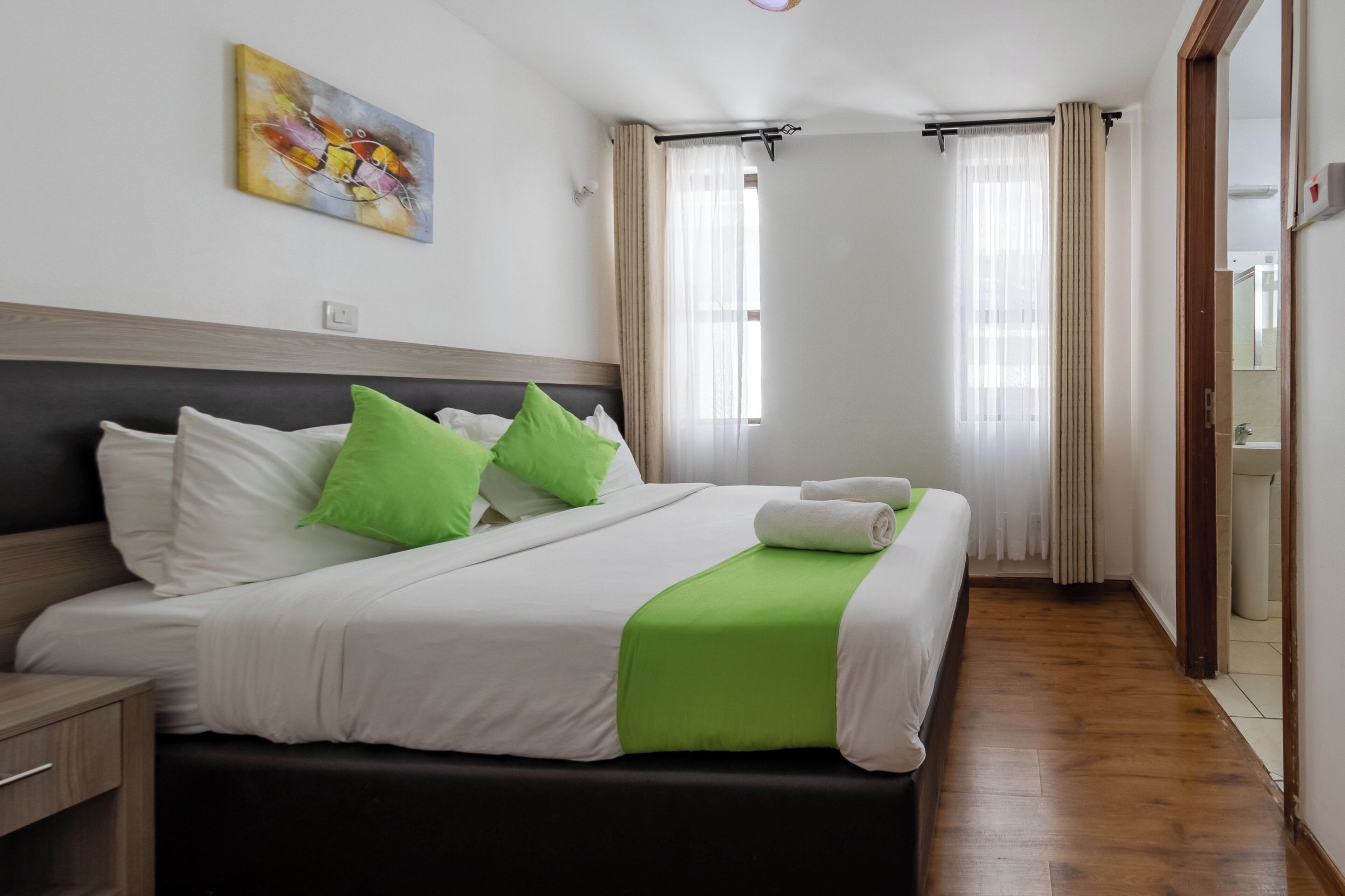 A neatly made bed with white and green bedding, a wooden headboard, and a painting on the wall.