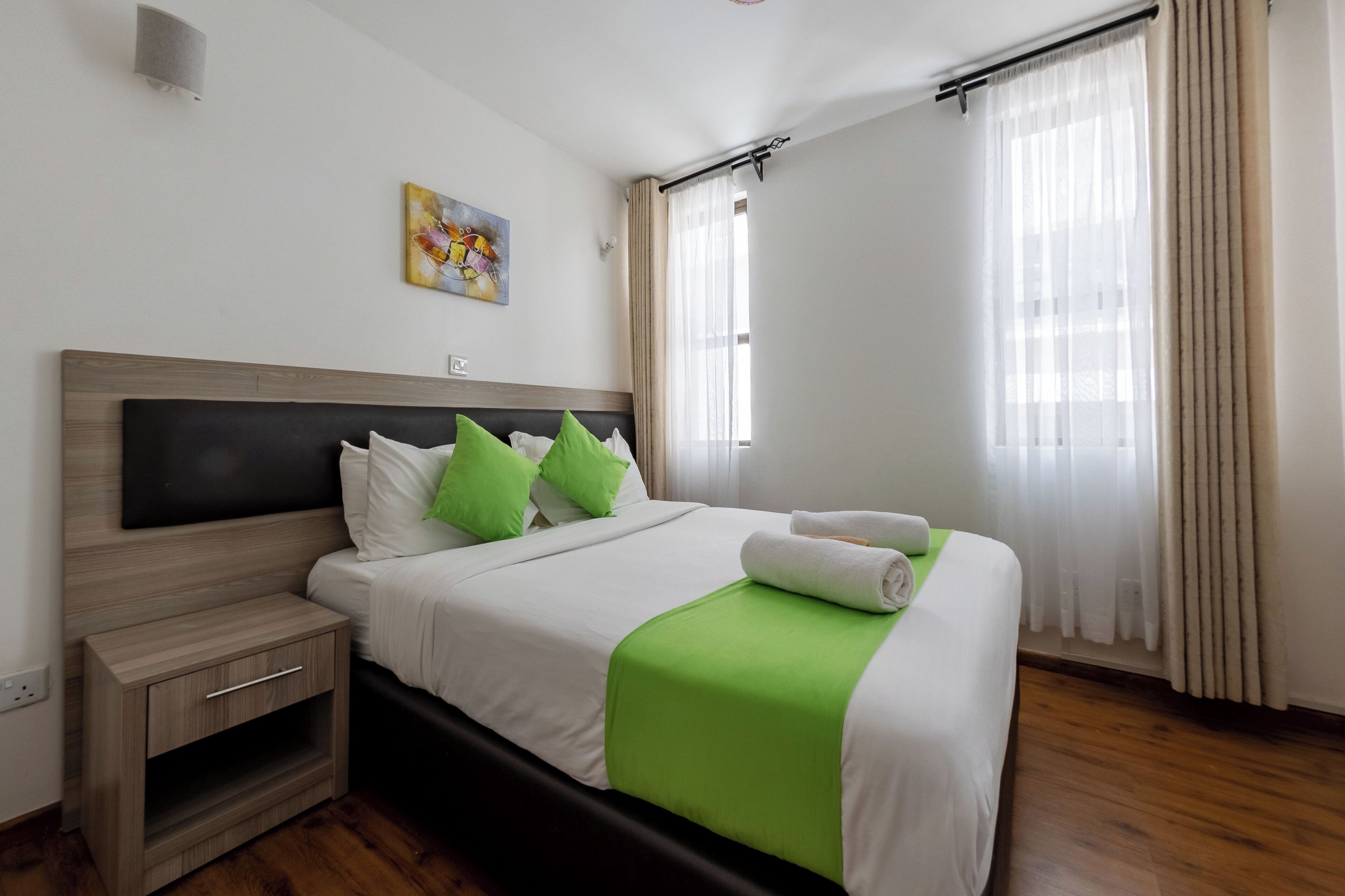 A neatly made bed with green pillows, a white blanket, and a rolled towel.