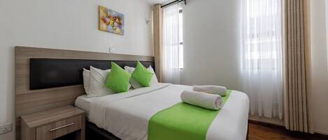 A neatly made bed with green pillows, a white blanket, and a rolled towel.