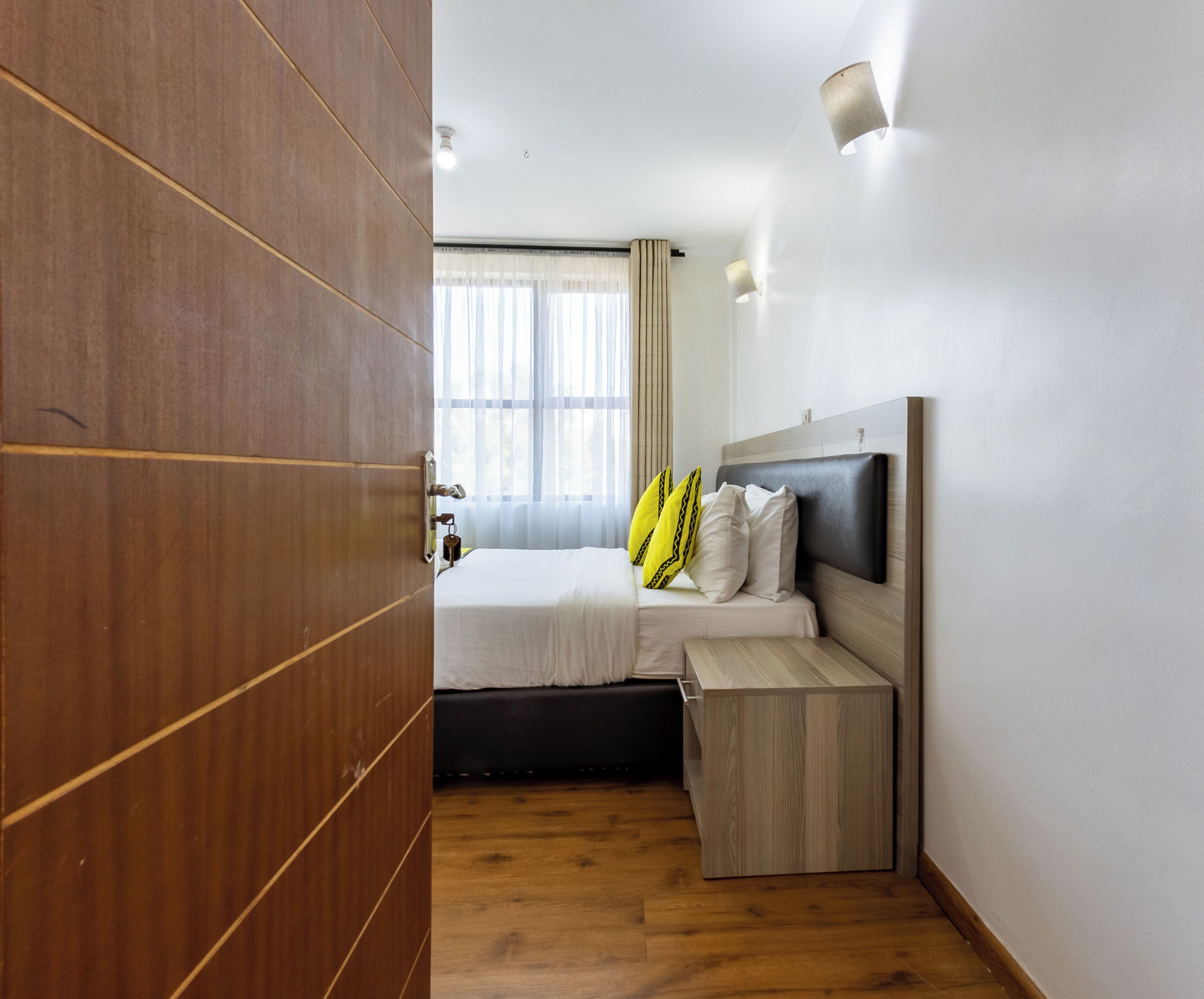 A hotel room with a wooden wardrobe, a bed with a headboard, a bedside table, and a window with curtains.