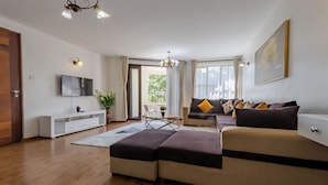 A modern living room with a sectional sofa, a white entertainment unit, a dining table with chairs, and a large window with curtains. - Erica Residences (Nairobi)