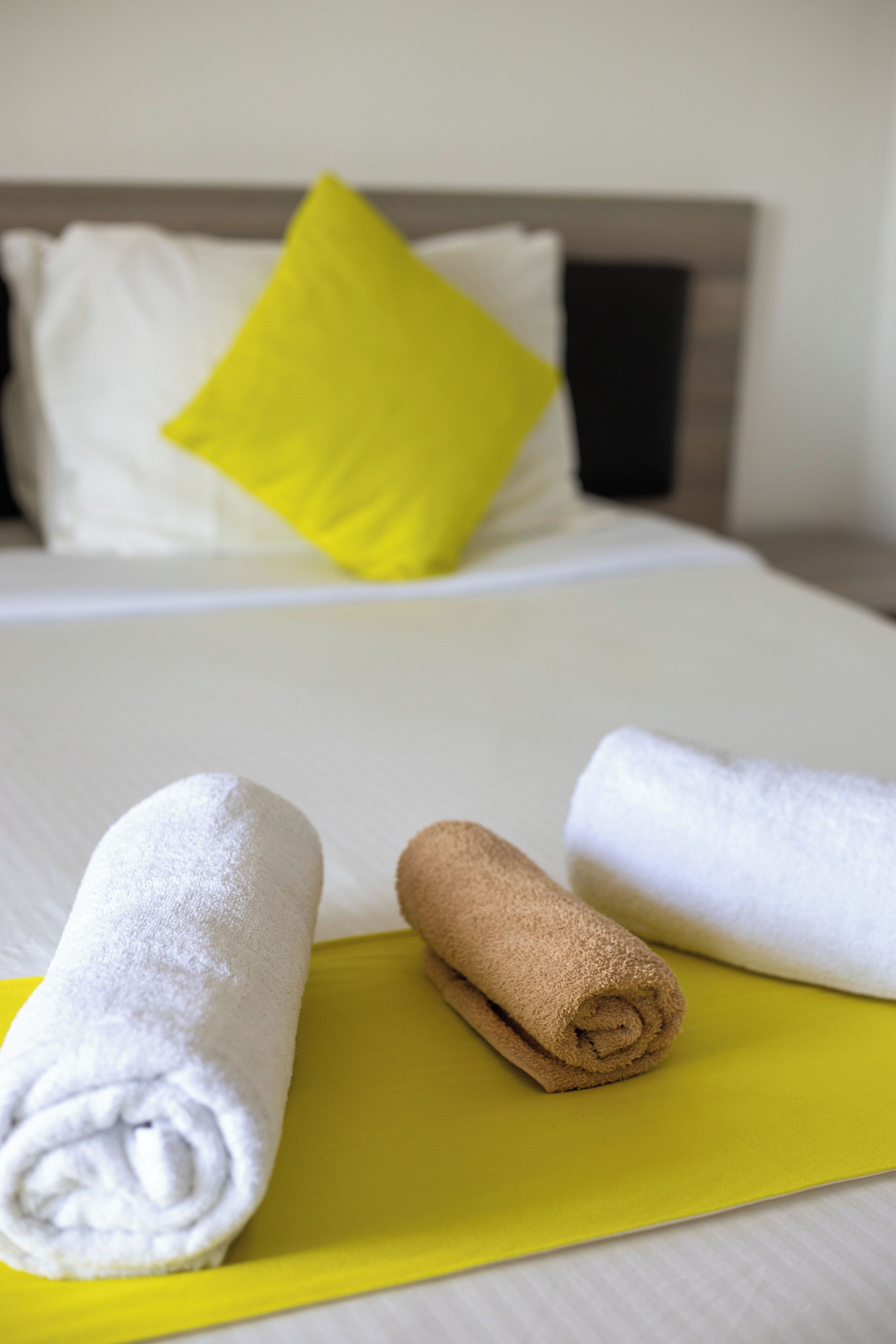 A neatly made bed with a bright yellow pillow and a white pillow, two rolled towels on a yellow mat, and a wooden headboard.