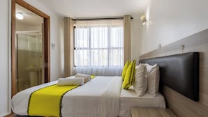 A hotel room with a bed, a bench, a window with curtains, and a door leading to a bathroom. - Erica Residences (Nairobi)