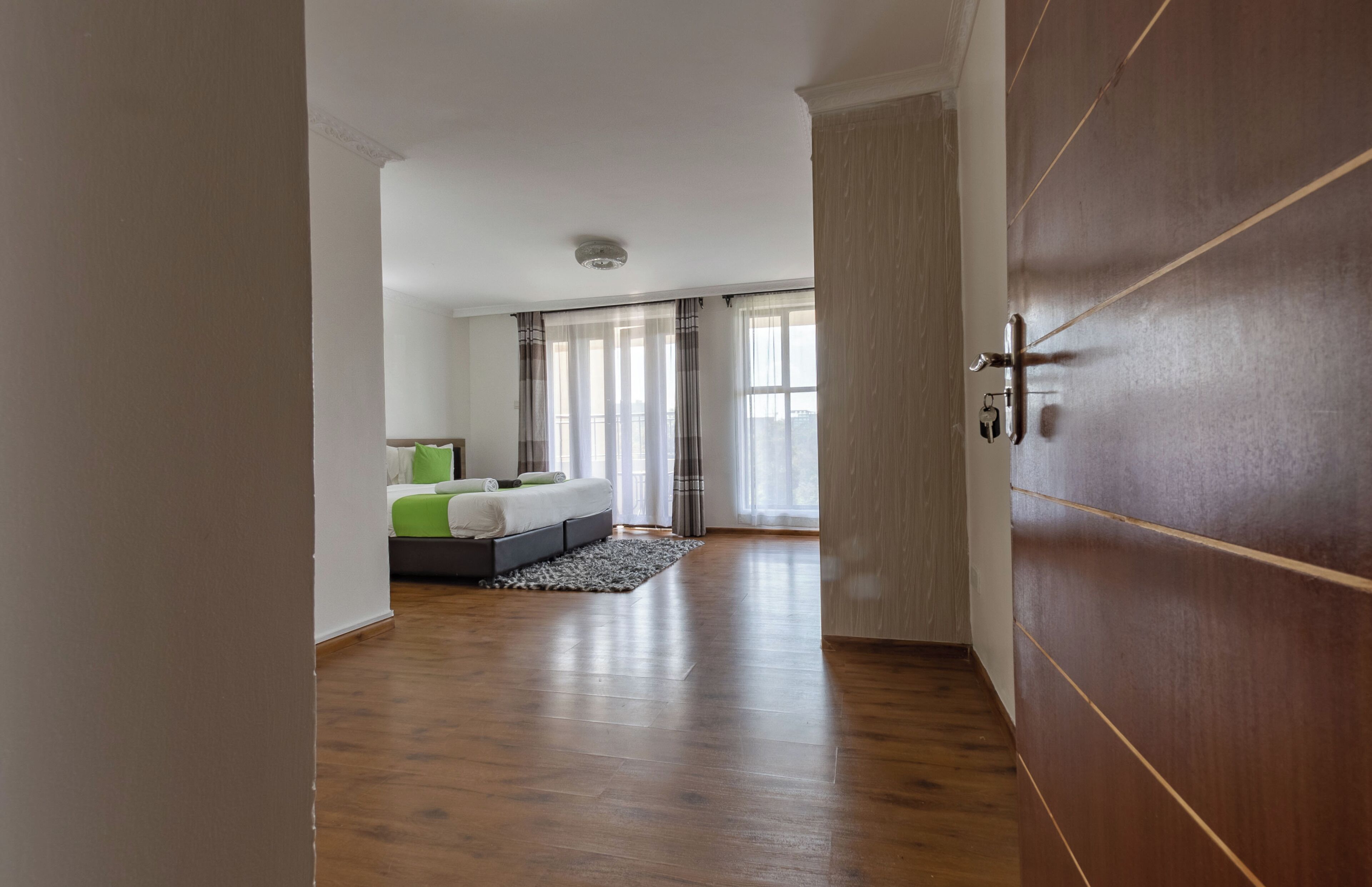 Une chambre moderne avec un plancher en bois, un lit recouvert d’une literie verte et blanche, et une vue sur la ville grâce à de grandes fenêtres.