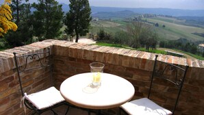 Une terrasse avec une table ronde et deux chaises, un verre à portée de main, et une vue sur des collines verdoyantes et des arbres.