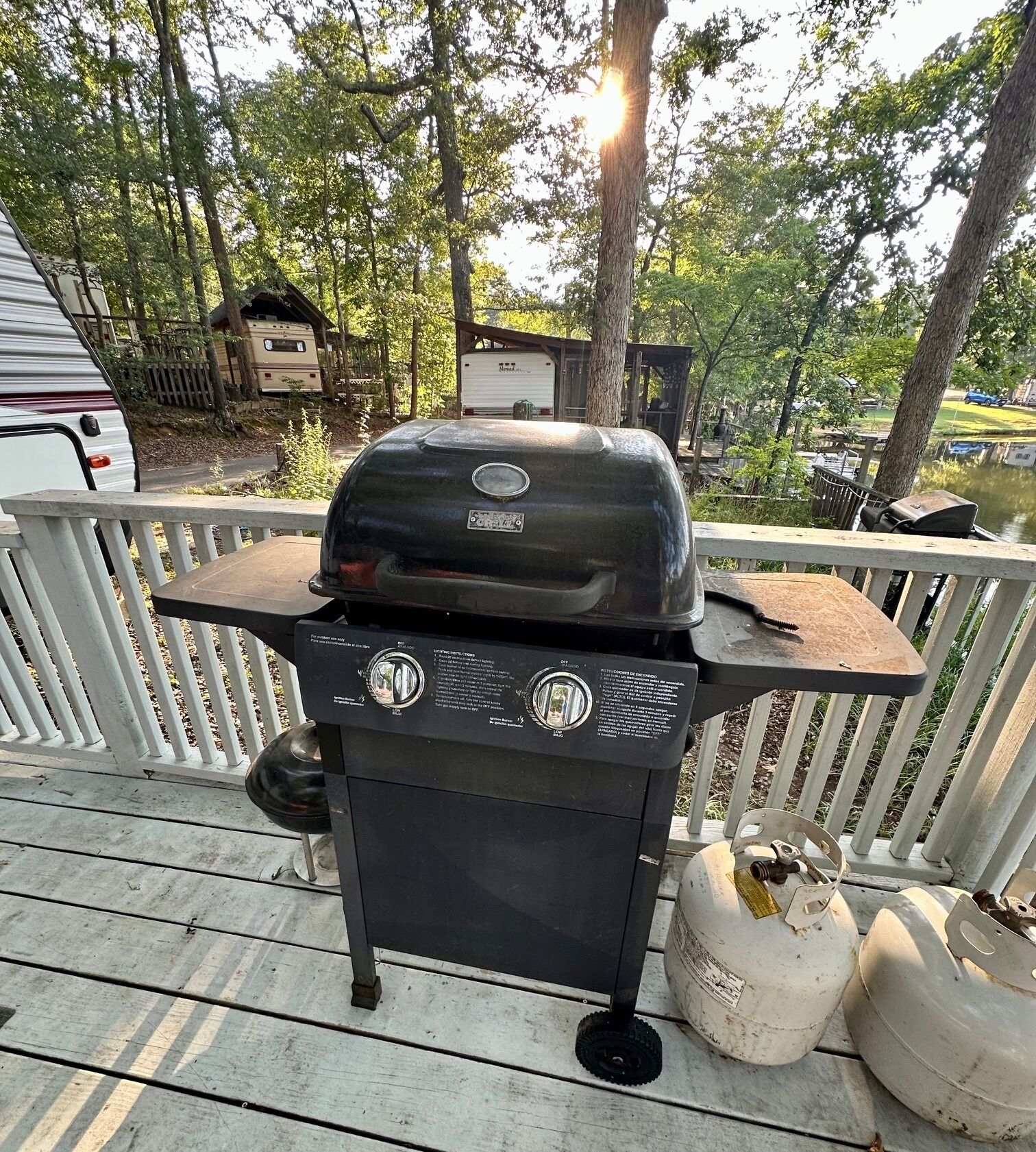 Ein tragbarer Grill auf einer Holzterrasse mit weißer Geländer, umgeben von Bäumen und Wohnhäusern im Hintergrund.