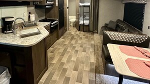 A compact RV kitchen with a sink, stove, and microwave. There is a dining area with a table and a couch. The RV has a tiled floor and a window with blinds. - Beautiful Lake Views at this Lake Front Camper on Private 25 Acre Lake (Mooresboro)