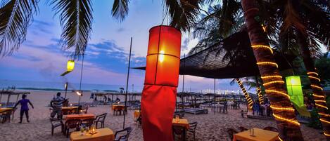 A beachside restaurant with outdoor seating, palm trees, and a sunset view.