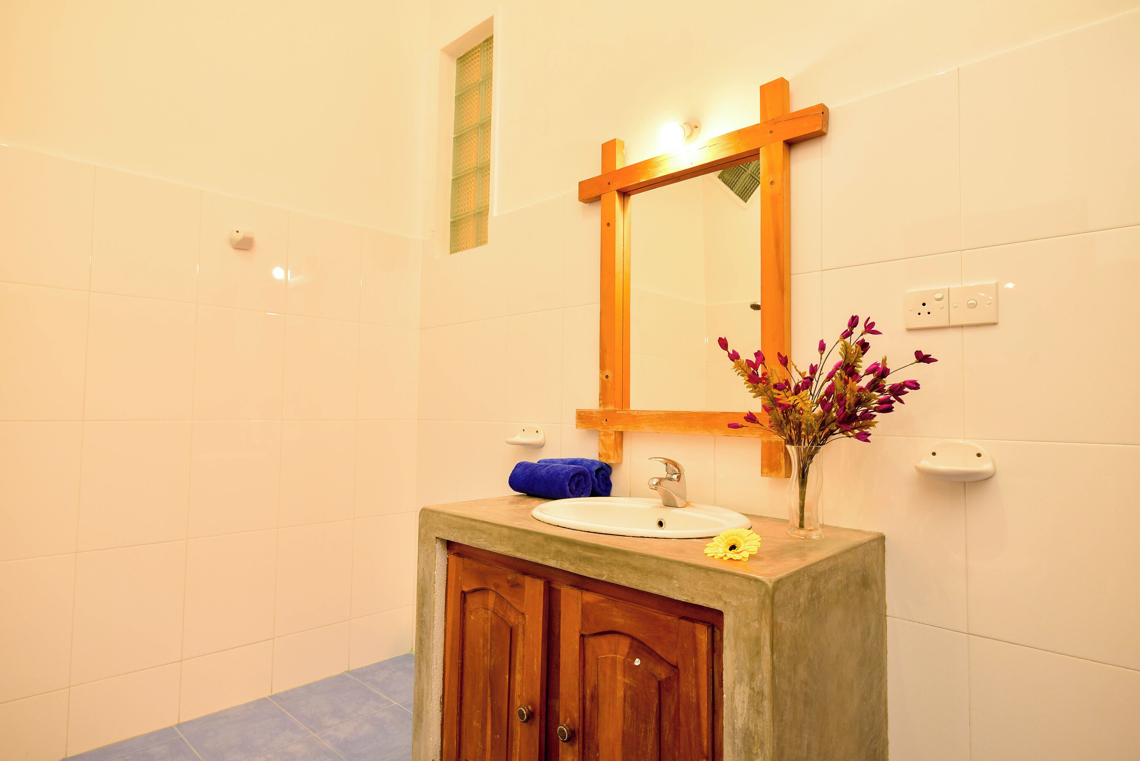 Une salle de bain avec un meuble-lavabo en bois, un lavabo blanc, un miroir avec un cadre en bois et un vase rempli de fleurs.
