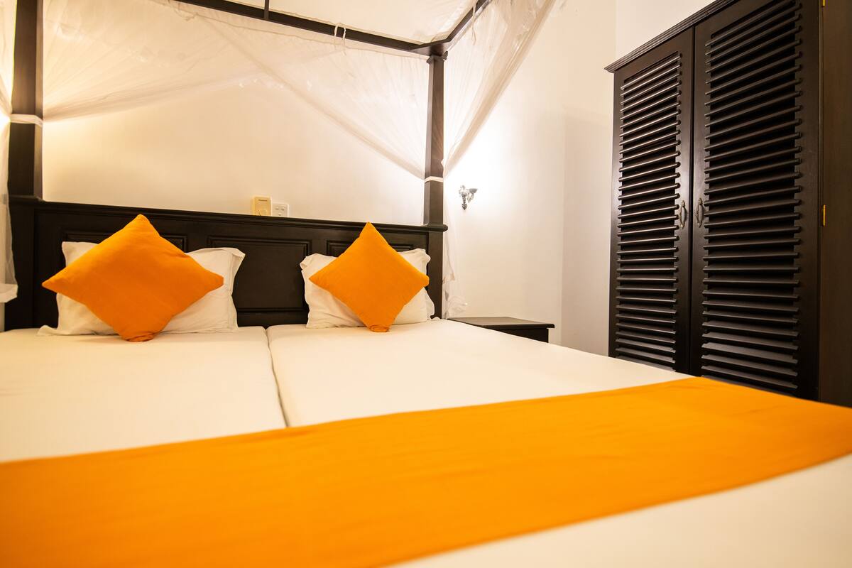 A hotel room with a bed, two orange pillows, a white bedspread, and a black wardrobe.