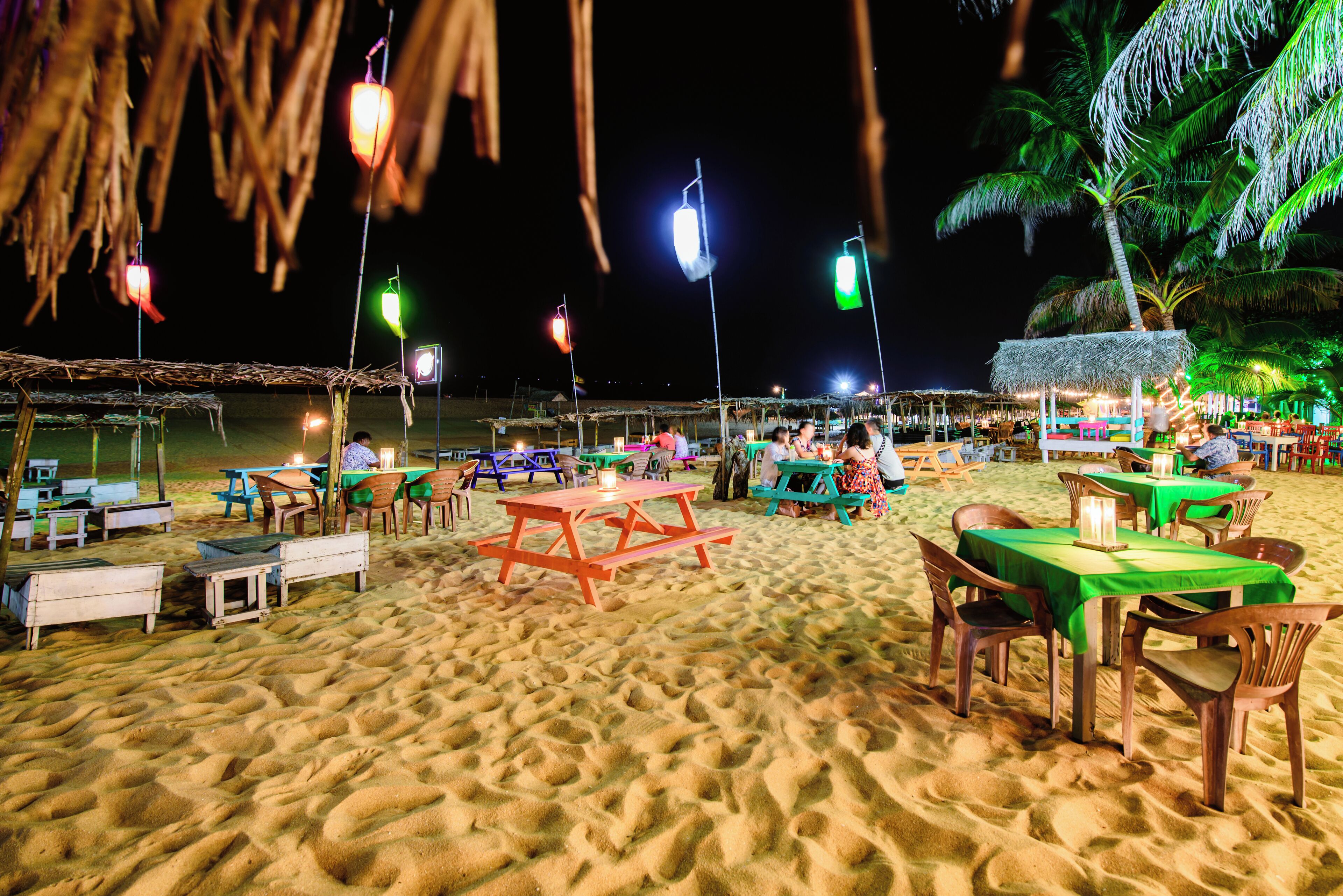 Een restaurant aan het strand met terras, tafels en stoelen, en een rieten dakconstructie op de achtergrond.