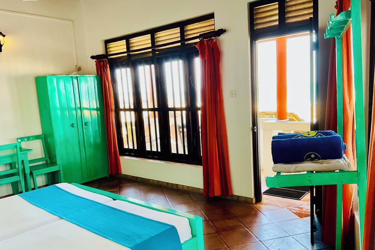 A room with a bed, a green chair, a wardrobe, a window with curtains, and a door with a towel rack.