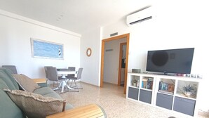 Un soggiorno con divano, tavolino, tavolo da pranzo con sedie, televisore su un mobile basso e un corridoio che porta in un'altra stanza.