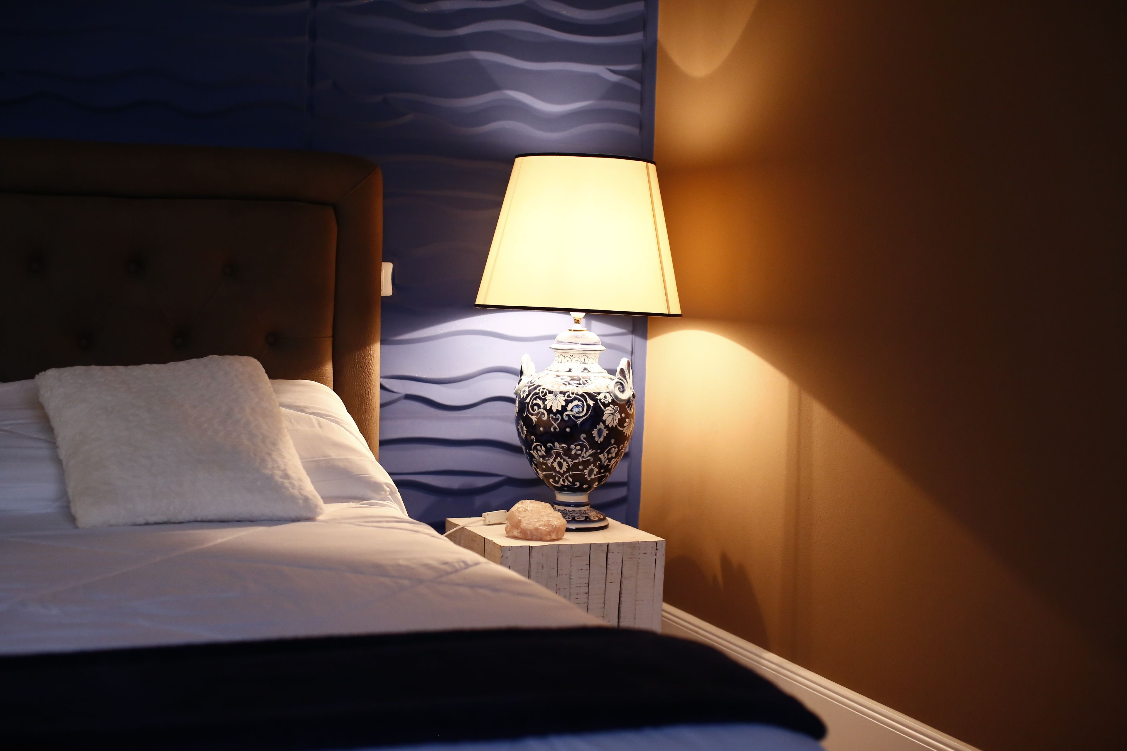 A neatly made bed with white linens and a fluffy pillow, a decorative lamp with a patterned shade on a wooden nightstand, and a textured blue wall in the background.