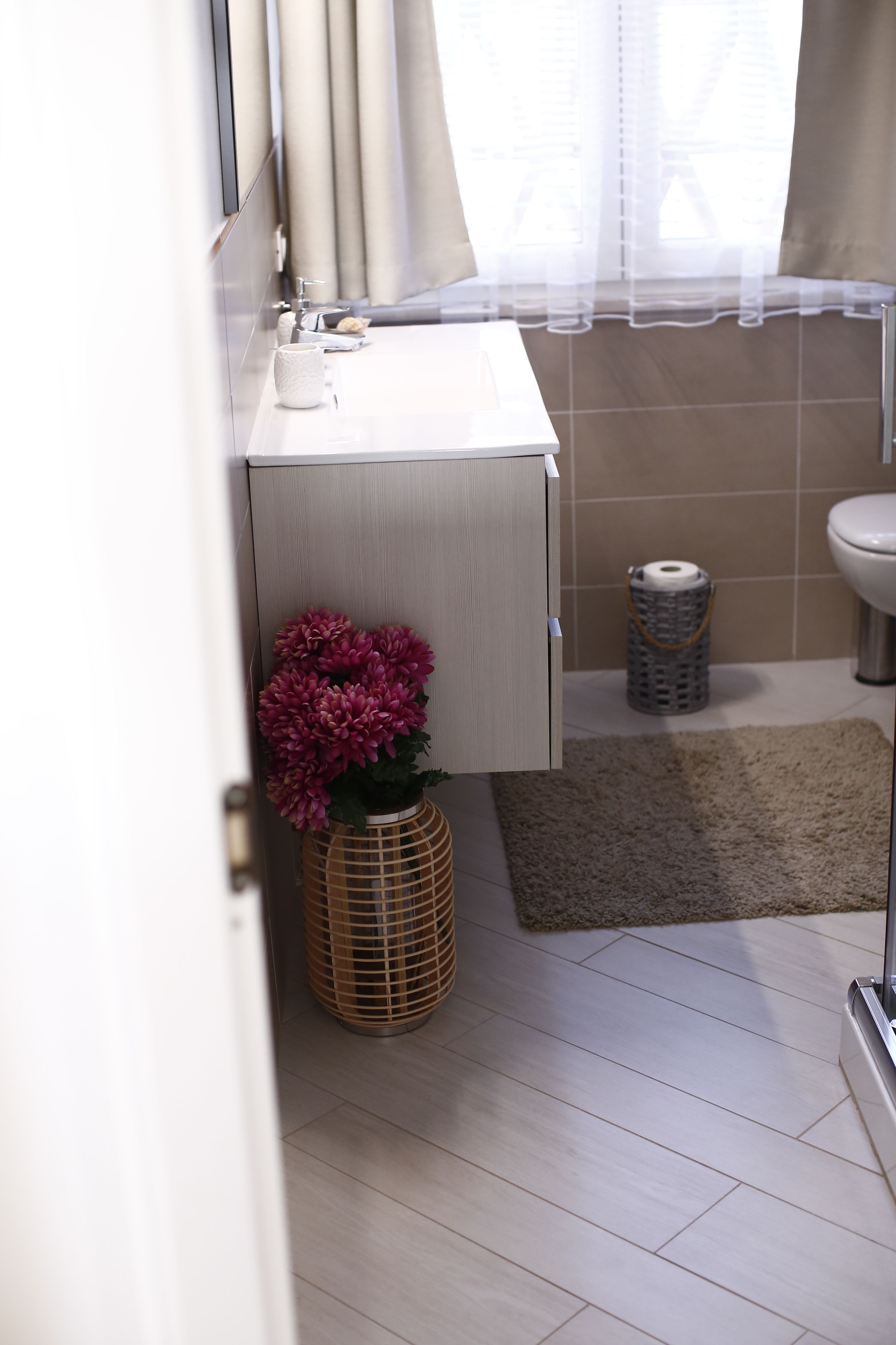 A modern bathroom with a white sink, mirror, and beige tiles. There is a wicker basket with a pink flower arrangement and a small rug on the floor.