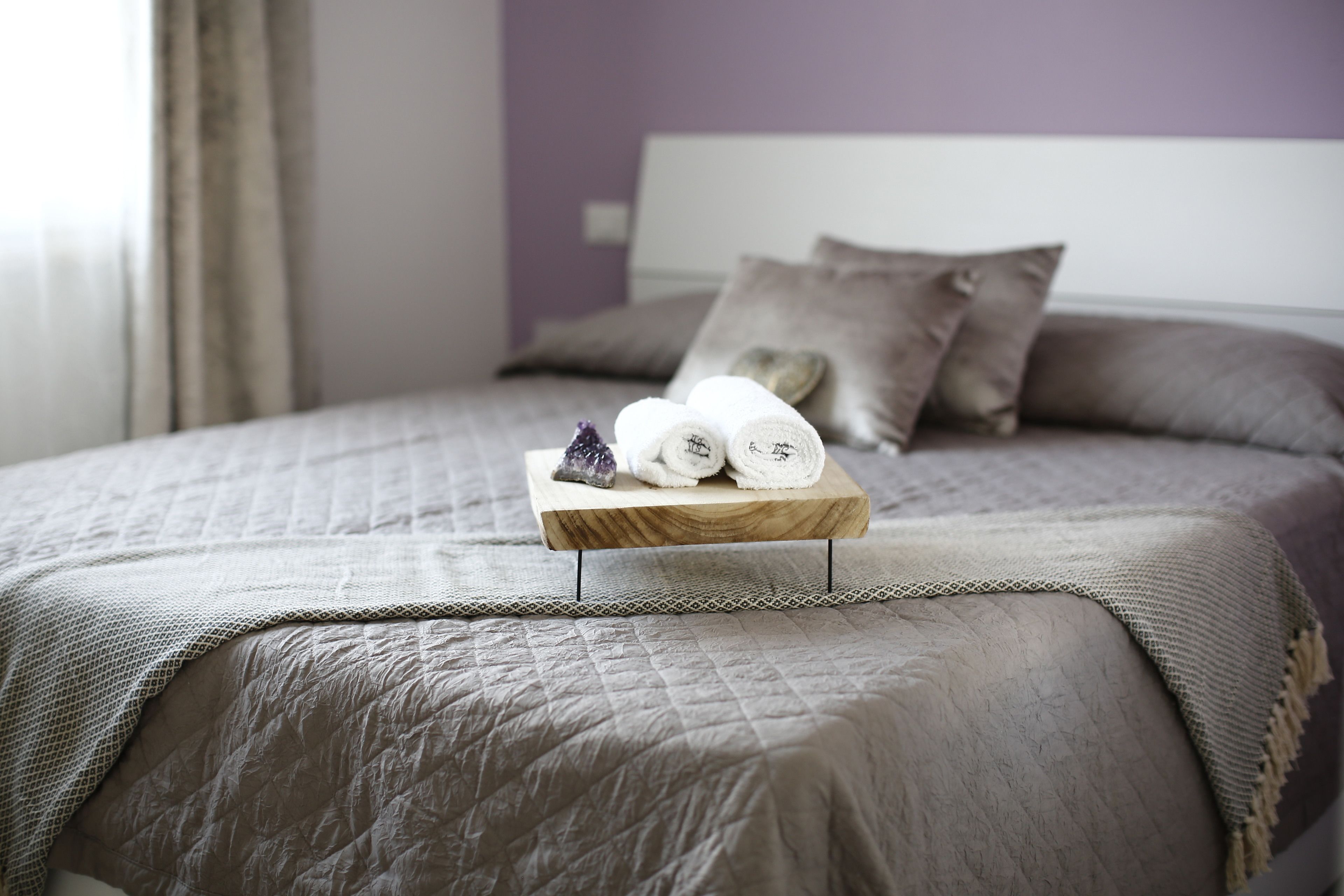 Ein ordentlich bezogenes Bett mit einer gesteppten Tagesdecke, ein Holztablett mit zusammengerollten Handtüchern und einem Kristallglas sowie ein Kissen mit einem herzförmigen Anhänger.