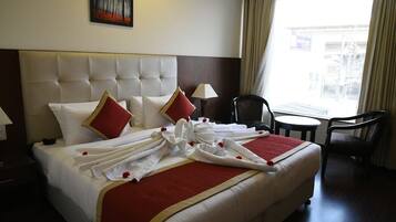 Quarto de hotel com uma cama grande, cabeceira, janela com cortinas, uma pequena mesa redonda e uma cadeira.