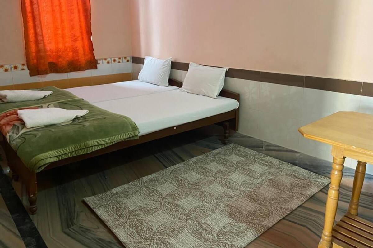 Una cama individual con colcha verde y almohadas blancas, una mesa de madera y una alfombra estampada sobre suelo de baldosas.