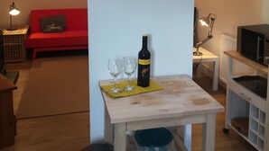 A living room with a red sofa, a wooden table with a bottle of wine and two glasses, a wall clock, and a small shelf.