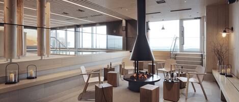 A modern interior with a central fire feature, wooden furniture, and large windows offering views.