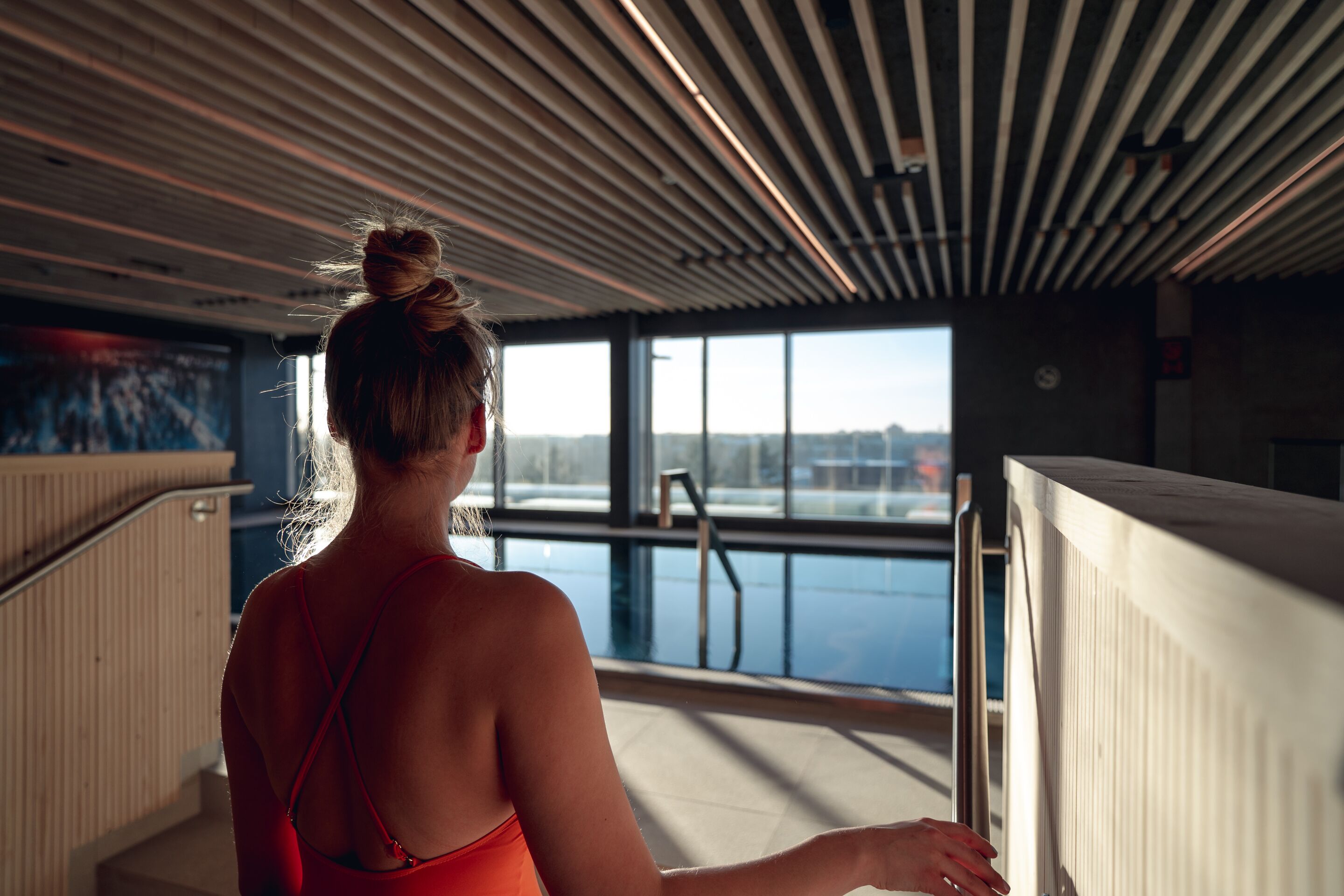 A woman in a red swimsuit standing by a pool with a view of a cityscape through large windows.