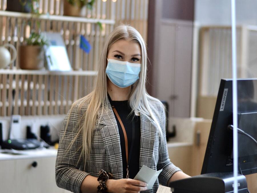 Una persona de pie en la recepción de una oficina moderna, con un monitor de computadora, una planta y un panel de pared de madera.