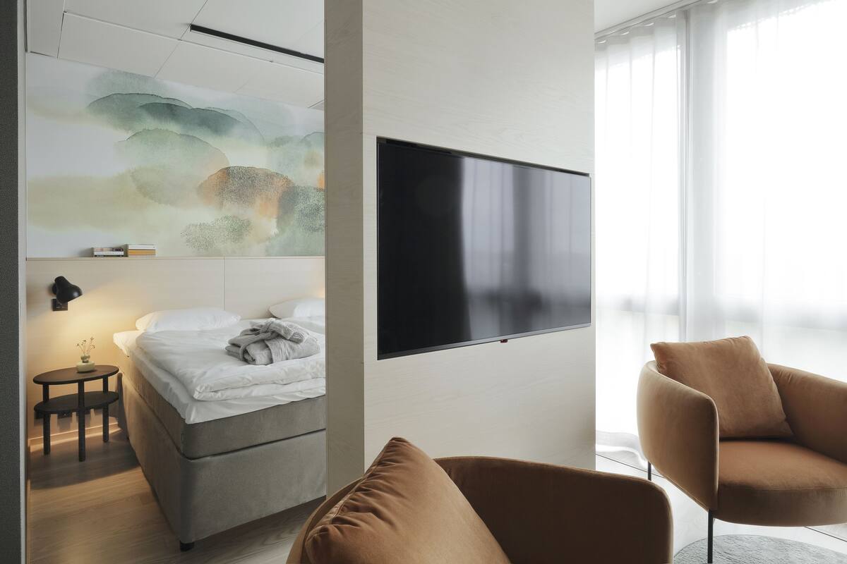A modern hotel room with a bed, a TV mounted on the wall, a small table with a lamp, and a large window with sheer curtains.