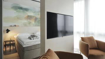 A modern hotel room with a bed, a TV mounted on the wall, a small table with a lamp, and a large window with sheer curtains.