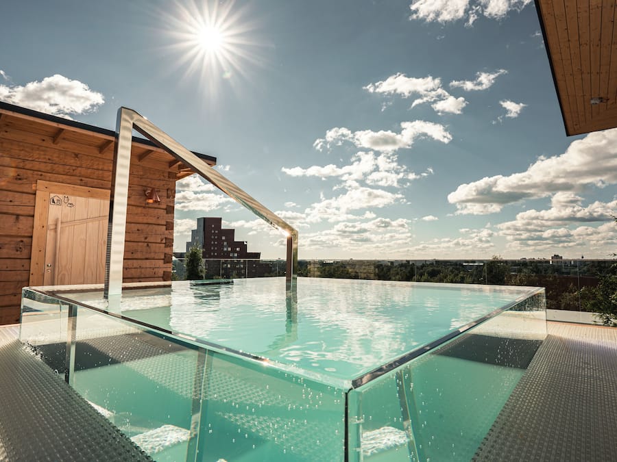 Una piscina d'hidromassatge infinita amb un solàrium de fusta i una construcció de cabana de fusta al fons.