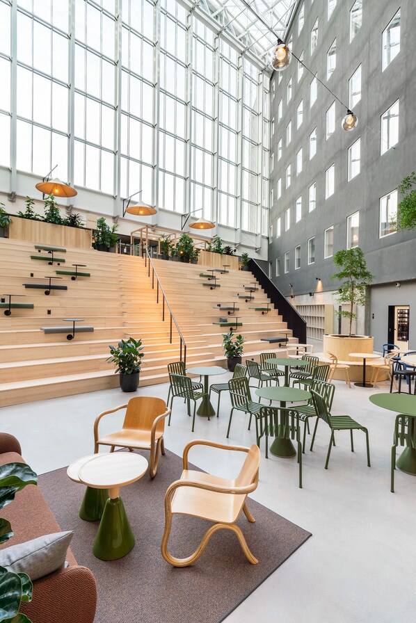 Een ruime binnenruimte met een hoge plafond, grote ramen en een trap die naar hogere verdiepingen leidt. Er zijn meerdere zithoeken met tafels en stoelen, en er staan potplanten door de hele ruimte.