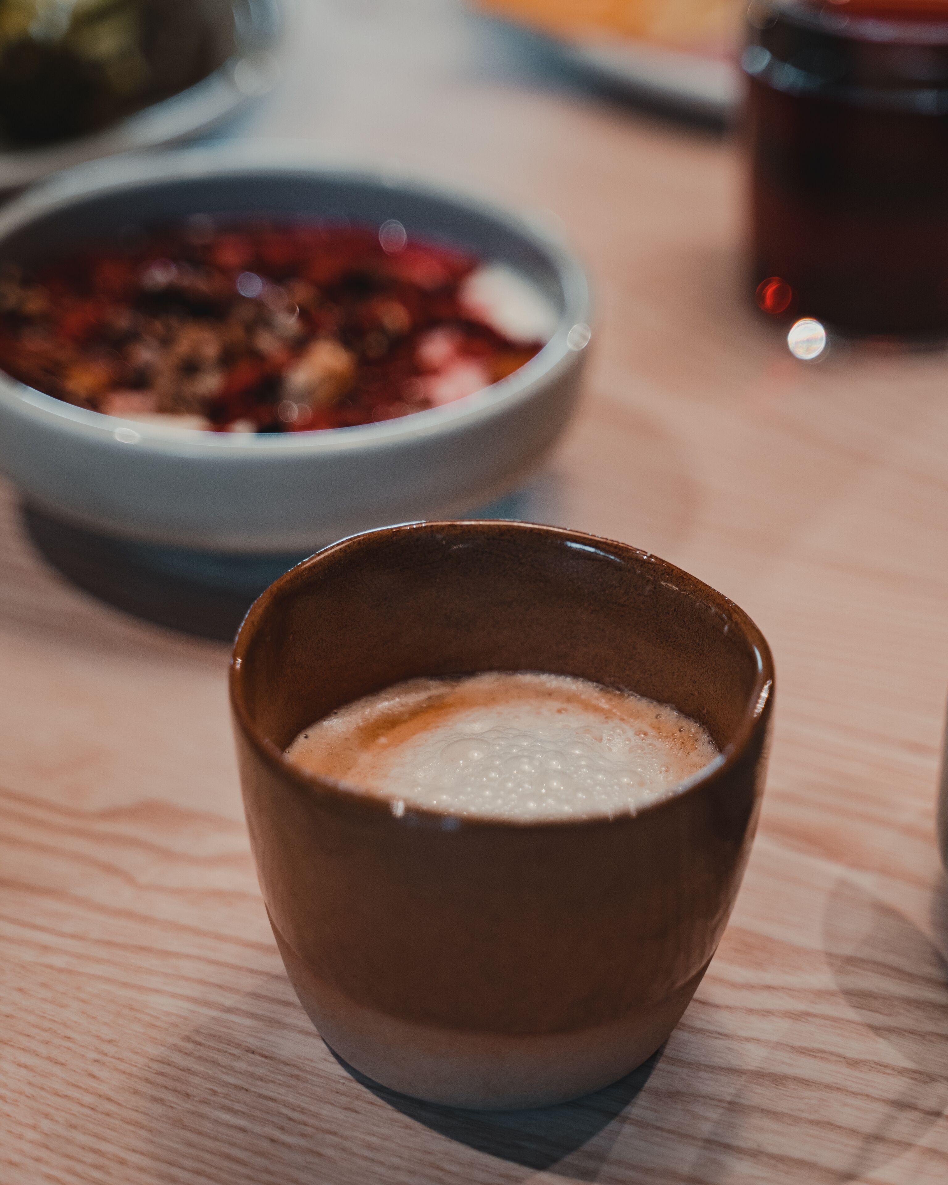Primer plano de una taza de cerámica con una bebida espumosa, un plato de comida al fondo y un vaso con líquido oscuro.