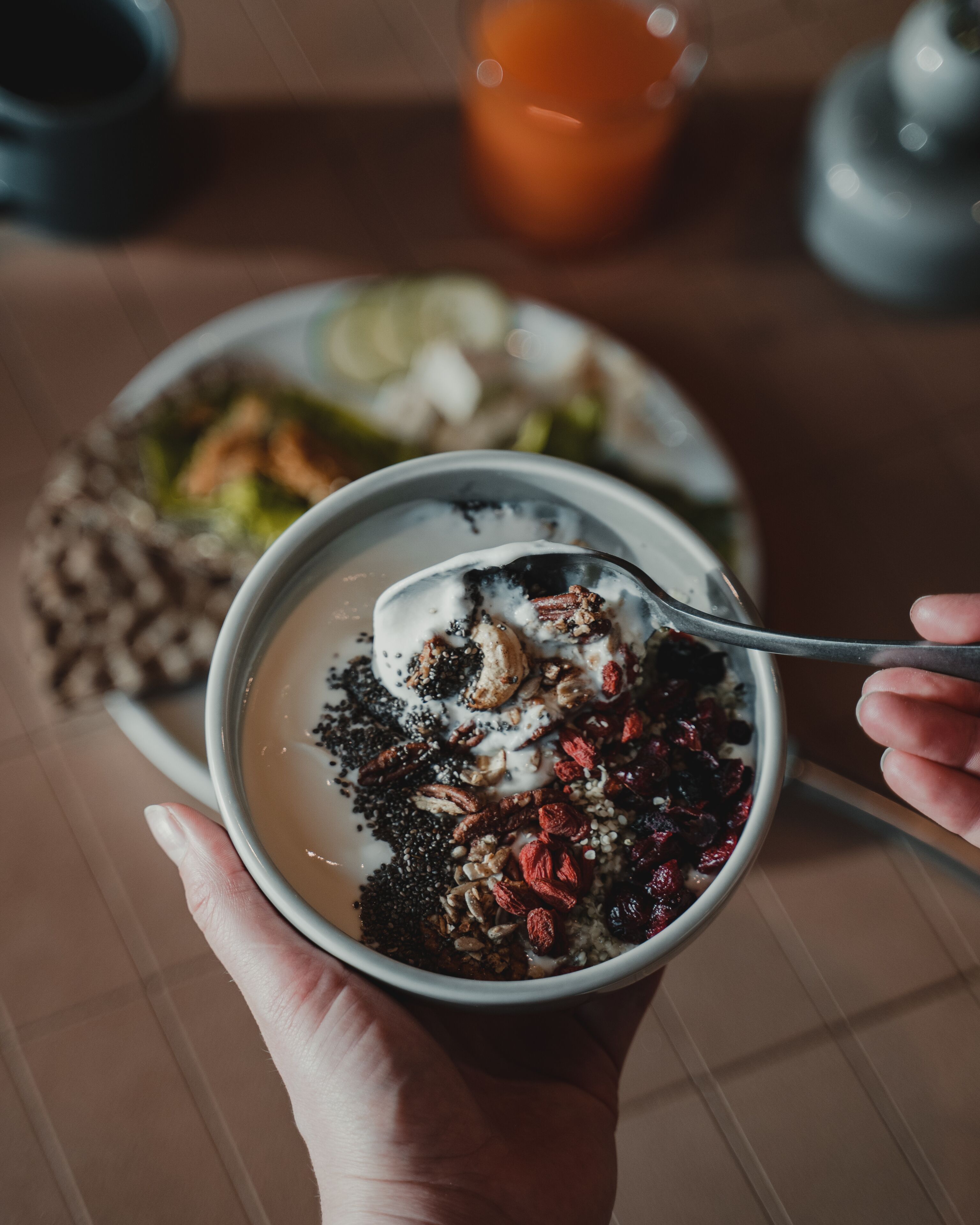 A person holding a bowl of oatmeal with toppings, a plate of food, and a glass of orange juice on a table.