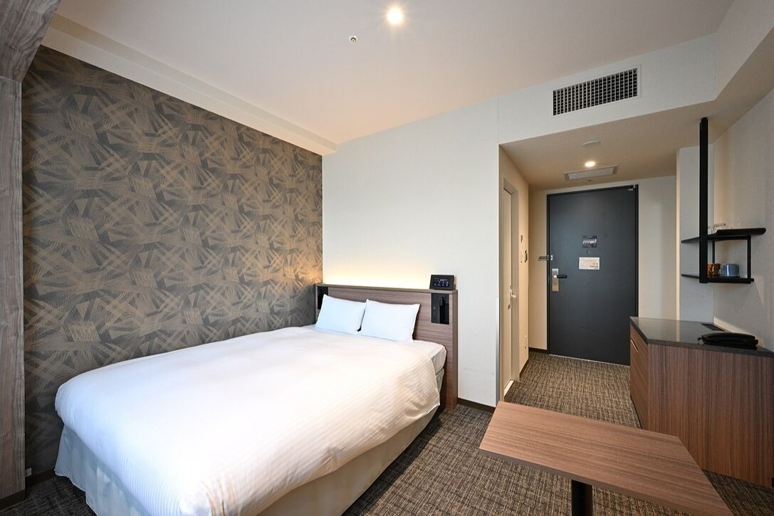A hotel room with a bed, a desk, a chair, a door, and a patterned wall.